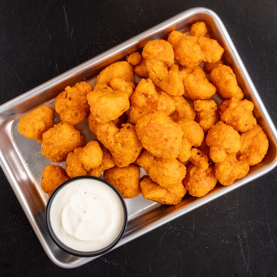 Golden-brown fried cauliflower bites in a metal tray with a side of creamy dipping sauce.