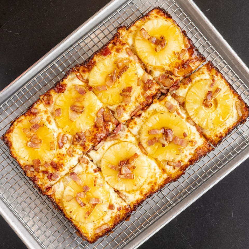 Detroit-style pizza with pineapple slices and bacon bits on a metal cooling rack.