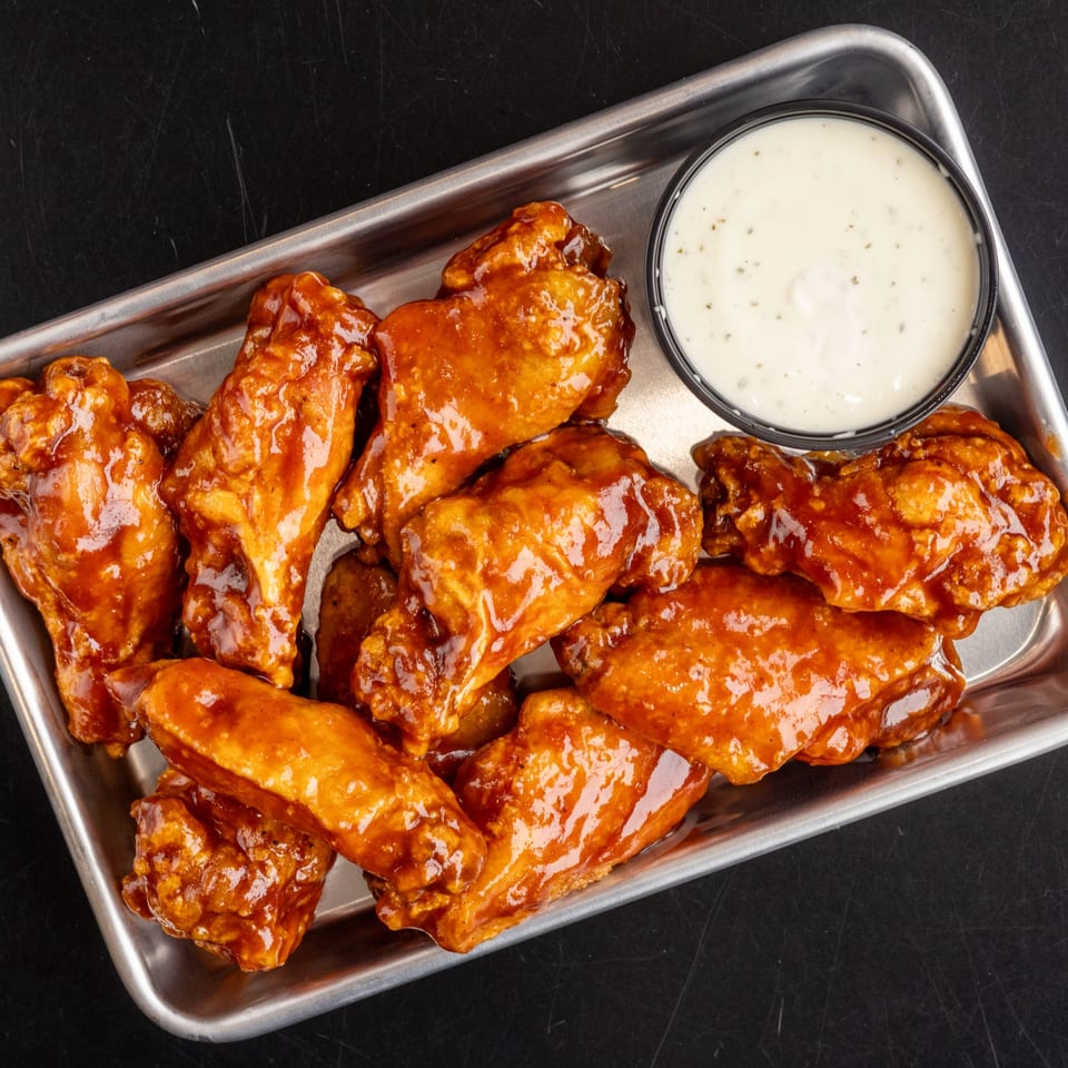 A metal tray filled with glossy, orange-glazed chicken wings and a side of creamy white dipping sauce.