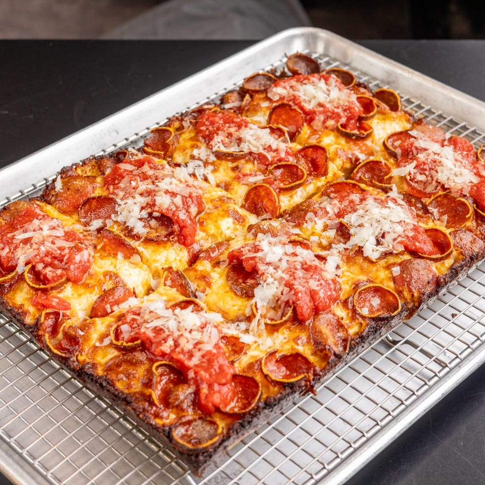 A rectangular Detroit-style pizza with pepperoni, cheese, and red sauce dollops on a cooling rack.